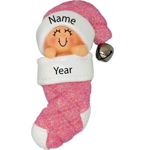 May include: Pink glitter stocking ornament with a baby doll inside wearing a white Santa hat with the word "Name" on it. The stocking has a white cuff with the word "Year" on it.