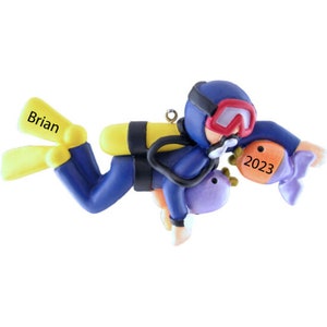 May include: A blue and yellow scuba diver ornament with a purple and orange fish. The diver has a yellow tank and a red face mask. The ornament is personalized with the name "Brian" and the year "2023".
