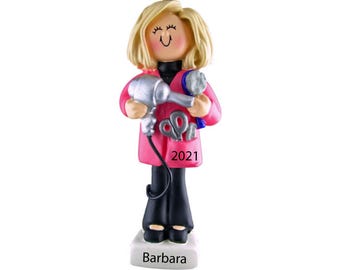 May include: A figurine of a hairstylist with blonde hair, wearing a pink coat and black pants. The figurine holds a silver hairdryer and a blue brush. The pocket of the coat has scissors and the year 2021. The base of the figurine says "Barbara".