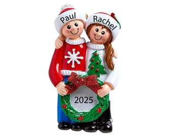 May include: A personalised Christmas bauble featuring a couple wearing winter jumpers and hats. The man is wearing a red jumper with a snowflake design and the woman is wearing a white jumper with a green Christmas tree design. They are holding a green wreath with a red bow and a white tag that says "2025". The bauble is decorated with glitter and has the names "Paul" and "Rachel" written on the hats.