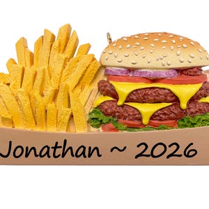 May include: A decorative ornament shaped like a double cheeseburger and a side of french fries in a tan container. The burger features sesame seeds, red onion, tomato, cheese, and lettuce. The text Jonathan ~ 2026 is printed on the front.