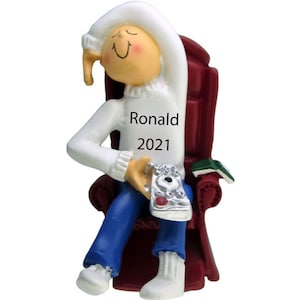 May include: A personalized Christmas ornament featuring a cartoon figure sitting in a red armchair, wearing a white sweater, blue jeans, and a white hat. The figure is holding a remote control and the ornament is inscribed with the name "Ronald" and the year "2021".