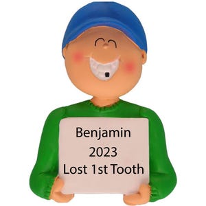May include: A green and blue personalized Christmas ornament of a boy with a missing tooth holding a sign that says "Benjamin 2023 Lost 1st Tooth".