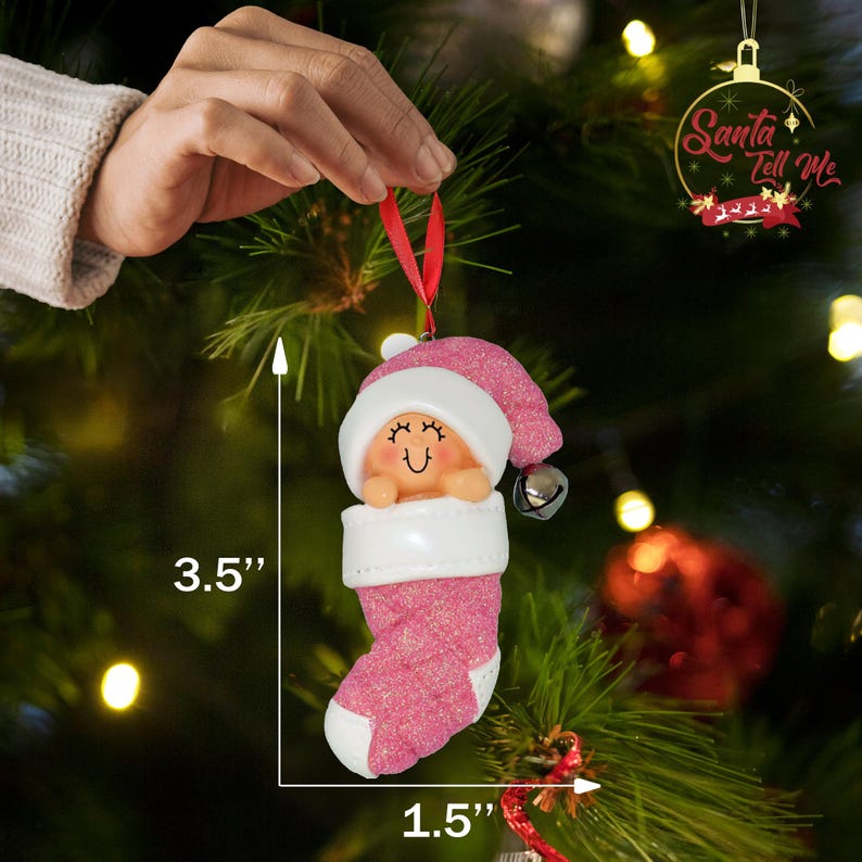 May include: Pink glitter stocking ornament with a white top and a smiling baby face. The ornament is 3.5 inches tall and 1.5 inches wide. It has a bell on the top and says "Santa Tell Me" on the tag.