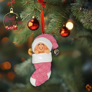 May include: Pink glitter stocking ornament with a baby face and a white hat. The ornament has a bell attached to the hat.