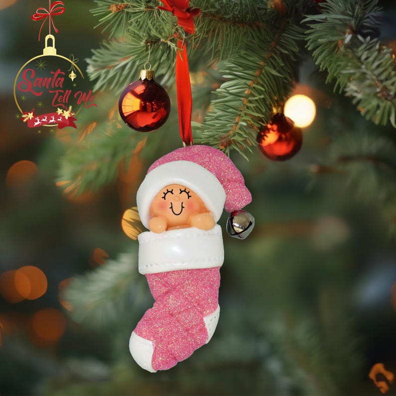 May include: Pink glitter stocking ornament with a baby face and a white hat. The ornament has a bell attached to the hat.