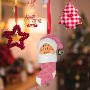 May include: Pink and white Christmas ornament of a baby in a stocking with a Santa hat. The ornament has a bell on the hat and a glittery finish.