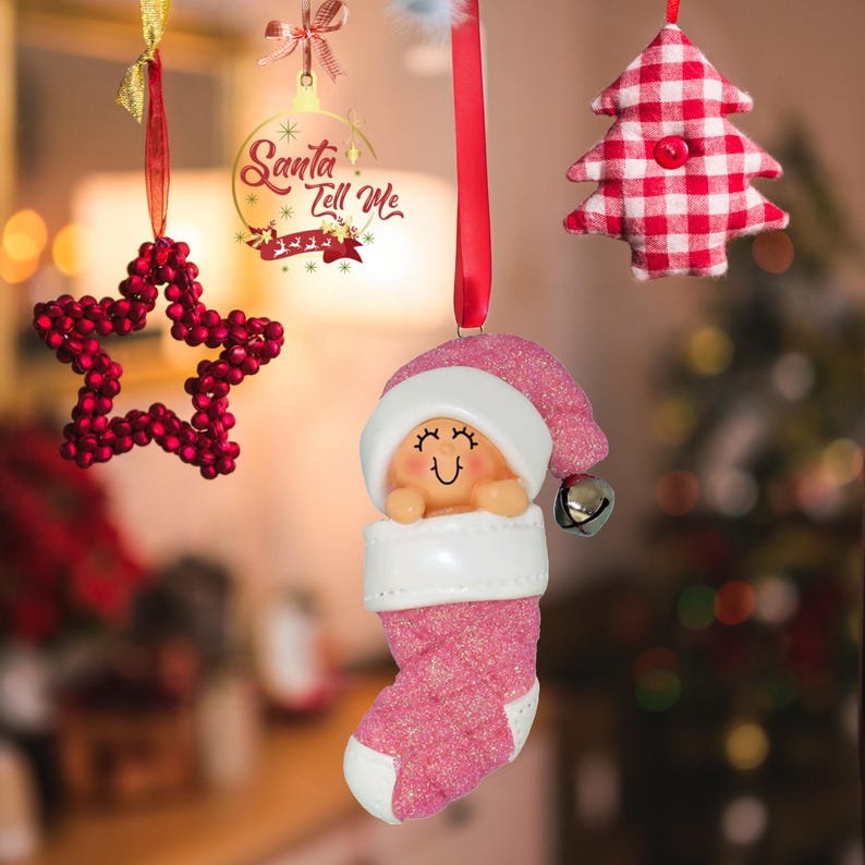 May include: Pink and white Christmas ornament of a baby in a stocking with a Santa hat. The ornament has a bell on the hat and a glittery finish.