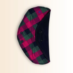 Puede incluir: Un babero de tela a cuadros negros y rojos con un respaldo de forro polar negro. El babero tiene tres broches negros en la parte posterior.