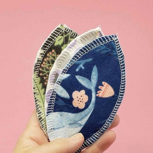 May include: Three reusable makeup remover pads in a hand. The pads are made of soft fabric and have a variety of floral patterns. The pads are blue, green, and white.