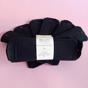 May include: A collection of dark blue reusable menstrual pads arranged in a fan shape. The pads have colored snaps and are labeled "Humes Reusable Menstrual Pad Mix". The label indicates the pads are for light, regular, and heavy flow.