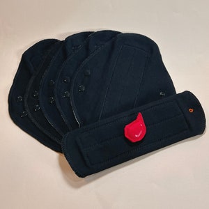 May include: A set of black reusable cloth menstrual pads with a red fabric drop symbol. The pads are folded and stacked on top of each other.