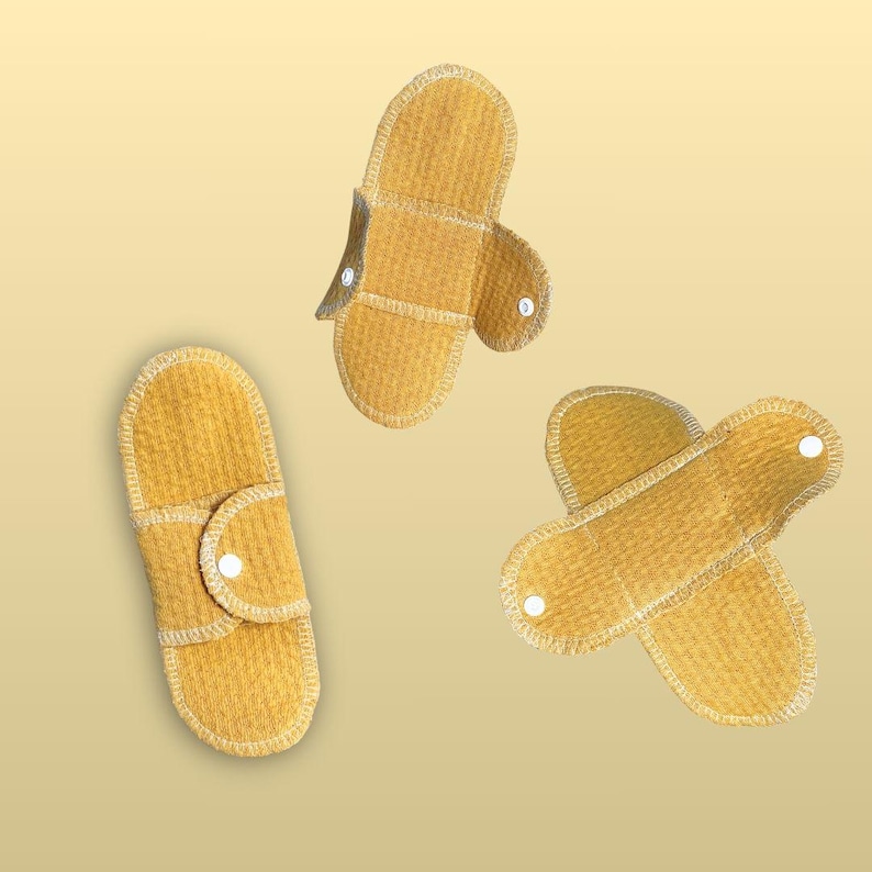 Three Golden Sand reusable cloth pads shown open, partly folded, and secured with snaps, displaying the pad shape, wings, organic cotton fabric, and stitching details.