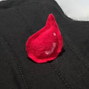 May include: A bright red felt teardrop shape on a black quilted fabric. The teardrop has a white highlight. The fabric is a dark color with visible stitching. The image is a close-up of a handmade item.
