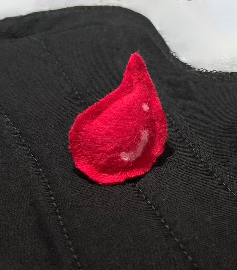 May include: A bright red felt teardrop shape on a black quilted fabric. The teardrop has a white highlight. The fabric is a dark color with visible stitching. The image is a close-up of a handmade item.