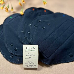 May include: A stack of navy blue reusable menstrual pads with snap closures. A small card reads "Humes Reusable Menstrual Pad Mix" and lists flow types: light, regular, and heavy. The pads have colored stitching.