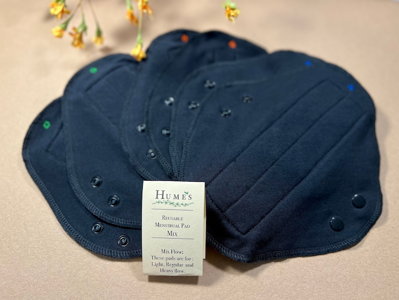 May include: A stack of navy blue reusable menstrual pads with snap closures. A small card reads "Humes Reusable Menstrual Pad Mix" and lists flow types: light, regular, and heavy. The pads have colored stitching.