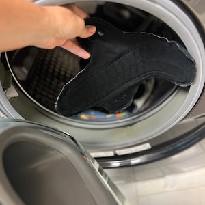 May include: A black reusable menstrual pad with a curved shape and white stitching is being placed into a washing machine. The pad has snaps for secure closure. The washing machine is gray.
