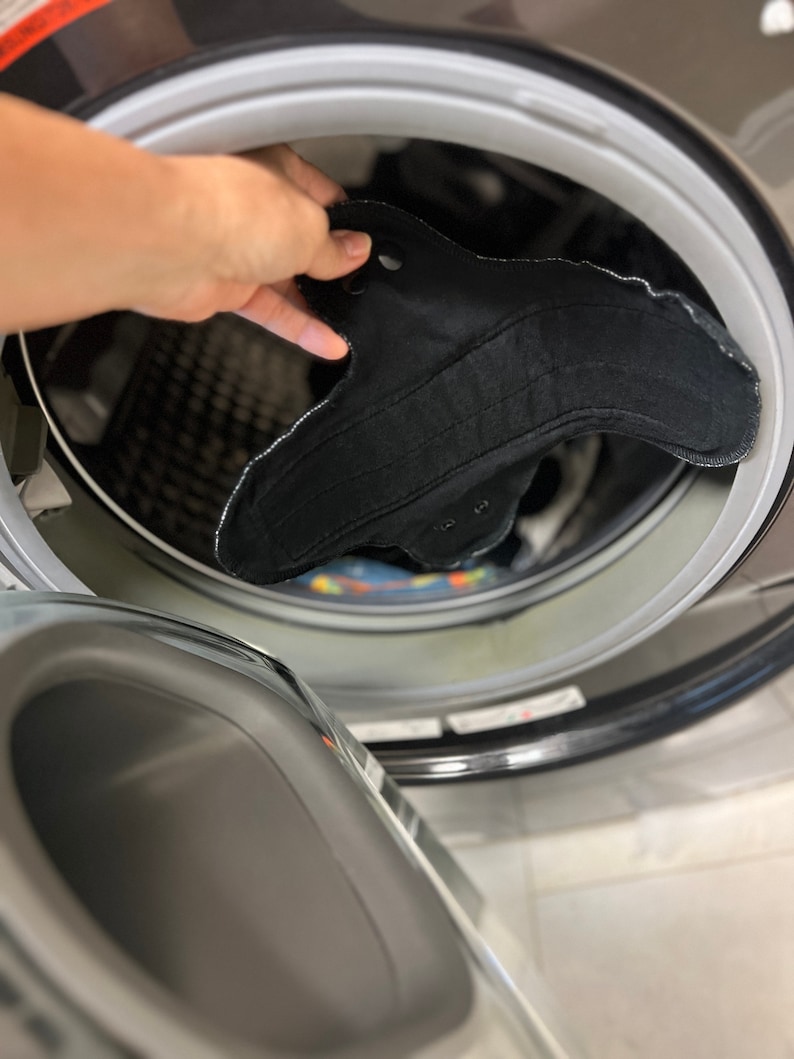 May include: A black reusable menstrual pad with a curved shape and white stitching is being placed into a washing machine. The pad has snaps for secure closure. The washing machine is gray.
