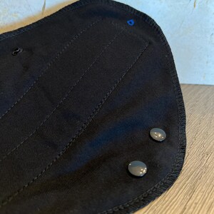 May include: Close-up of a black fabric item with a curved shape. The item has two gray snap closures and a blue stitch. The fabric has vertical stitched lines and a stitched border.