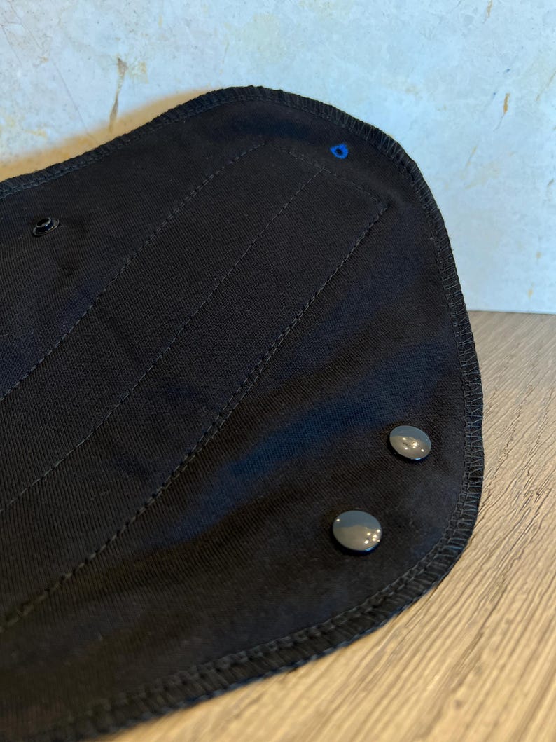 May include: Close-up of a black fabric item with a curved shape. The item has two gray snap closures and a blue stitch. The fabric has vertical stitched lines and a stitched border.
