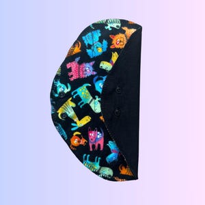 Puede incluir: Una compresa menstrual reutilizable negra con un estampado colorido de perros de dibujos animados. La compresa tiene un respaldo de tela negra y broches a los lados.