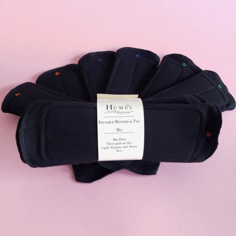 May include: A collection of dark blue reusable menstrual pads arranged in a fan shape. The pads have colored snaps and are labeled "Humes Reusable Menstrual Pad Mix". The label indicates the pads are for light, regular, and heavy flow.