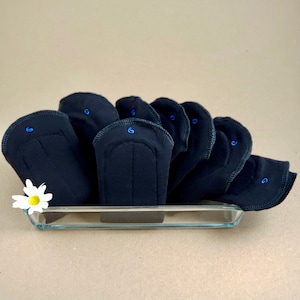 May include: A set of black reusable cloth menstrual pads in a glass dish. The pads are folded and stacked on top of each other. A small white daisy is in the foreground.