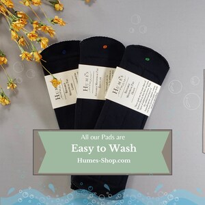 May include: Three black reusable menstrual pads with white labels, each with a different colored dot. The pads are labeled "Liners", "Regular", and "Heavy". Text on the image reads "All our Pads are Easy to Wash" and "Humes-Shop.com".
