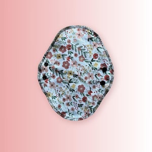 Puede incluir: Una compresa menstrual de tela blanca con estampado floral en tonos rosa, rojo y amarillo. La compresa tiene un borde cosido negro y dos botones negros.
