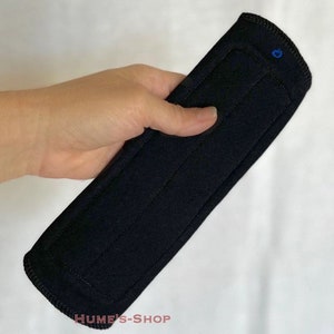 May include: A black, rectangular fabric item with stitched lines and a rounded end. A small blue button is visible on the top. The text "HUME'S-SHOP" is printed at the bottom. The item is held in a hand.