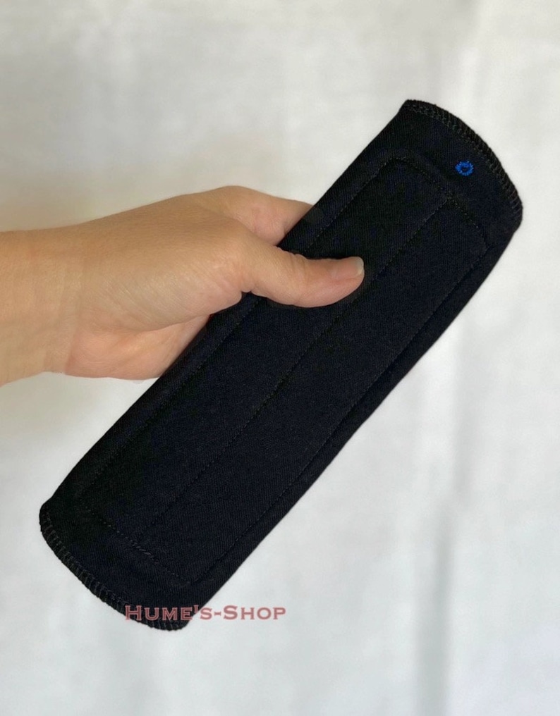 May include: A black, rectangular fabric item with stitched lines and a rounded end. A small blue button is visible on the top. The text "HUME'S-SHOP" is printed at the bottom. The item is held in a hand.