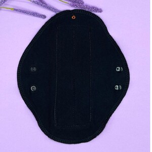 May include: A black reusable menstrual pad with snap closures. The pad has a contoured shape and is made of a soft, absorbent material. The pad is shown on a light purple background with purple decorative sprigs.
