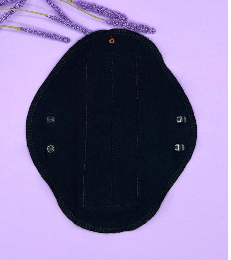 May include: A black reusable menstrual pad with snap closures. The pad has a contoured shape and is made of a soft, absorbent material. The pad is shown on a light purple background with purple decorative sprigs.
