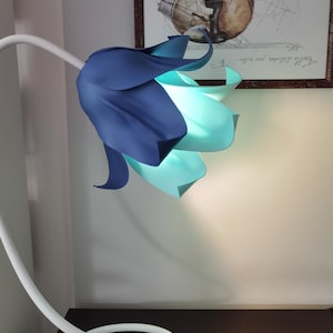 May include: A unique, flower-shaped lamp with a white, curved stem and base. The lamp shade is a layered design in shades of blue and teal, emitting a soft glow. The lamp is on a dark brown surface, with a framed artwork in the background.