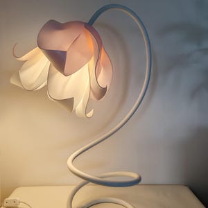 以下が含まれることがあります： 白い花の形をしたシェードが付いたテーブルランプ。ランプは白い柔軟なステムを持ち、ベースに巻き付けられています。