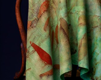 May include: A green and brown textile with a leaf pattern draped over a wooden chair. The fabric has a watercolour design with green, brown, and red shades. The textile's texture appears soft and flowing, with a natural, organic aesthetic.