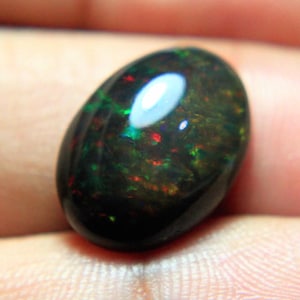 May include: A polished, oval-shaped black opal gemstone. The opal displays a play of color with flashes of red, green, and yellow. The stone is smooth and reflective, showcasing a natural, iridescent pattern.
