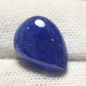 May include: A polished, teardrop-shaped gemstone in a deep, saturated blue hue. The stone has a smooth, glossy surface with a slight sheen, and is set against a white, textured background. The gem is likely a cabochon.