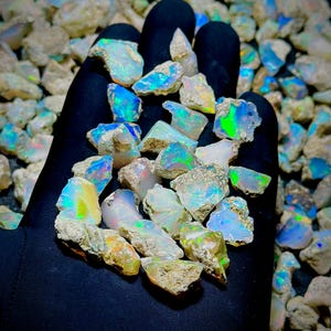 Può includere: Una collezione di pietre di opale grezze e non lucidate con una base bianco latte e vivaci bagliori di luce iridescente blu, verde e arancione. Le pietre presentano forme irregolari e frastagliate con superfici ruvide e testurizzate.