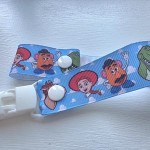 May include: A light blue ribbon dummy clip featuring a repeating pattern of cartoon characters including a cowgirl, a potato-shaped figure, and a green dinosaur against a sky background with white clouds, finished with a white plastic clip.