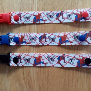 May include: Three baby pacifier clips featuring a Spider-Man design. Each clip has a different coloured plastic clip: red, blue, and black. The white ribbon displays Spider-Man and spiderweb graphics in red, blue, and black.