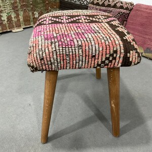 May include: A small, square stool with a woven top featuring a pattern of pink, white, and brown. The stool has three wooden legs. The legs are light brown and slightly angled.