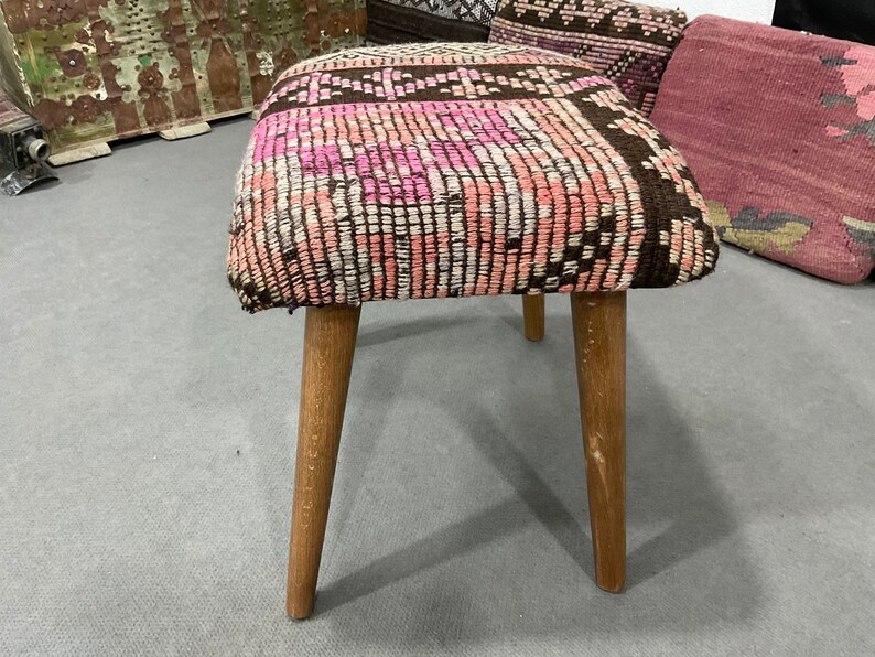May include: A small, square stool with a woven top featuring a pattern of pink, white, and brown. The stool has three wooden legs. The legs are light brown and slightly angled.