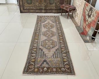 May include: A long, rectangular runner rug featuring a central beige field with three repeating geometric hexagonal medallions in shades of brown, gold, and gray. The border displays intricate floral and geometric motifs in dark brown and muted tones.