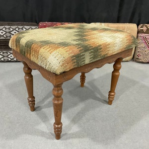 Può includere: Uno sgabello in legno con un cuscino per sedile in tappezzeria tessuta. Il cuscino presenta un motivo geometrico in tonalità di marrone, verde e arancio.