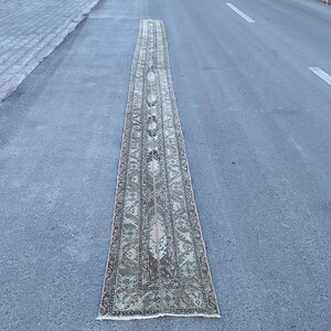 Puede incluir: Una alfombra larga y rectangular con un intrincado patrón de diseños florales y geométricos beige, marrón y verde. La alfombra está colocada sobre una superficie de asfalto gris, mostrando su longitud y detallada artesanía. Los bordes de la alfombra están acabados con una sutil franja.