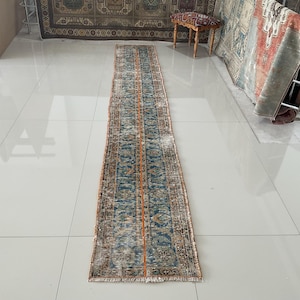 May include: A long, rectangular rug with a blue, orange, and white geometric pattern. The rug has a fringe border and is laid out on a white tiled floor.
