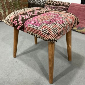 May include: A small, square stool with a patterned top in shades of pink, brown, and cream. The seat is supported by four light-colored wooden legs. The stool has a vintage or handmade aesthetic.