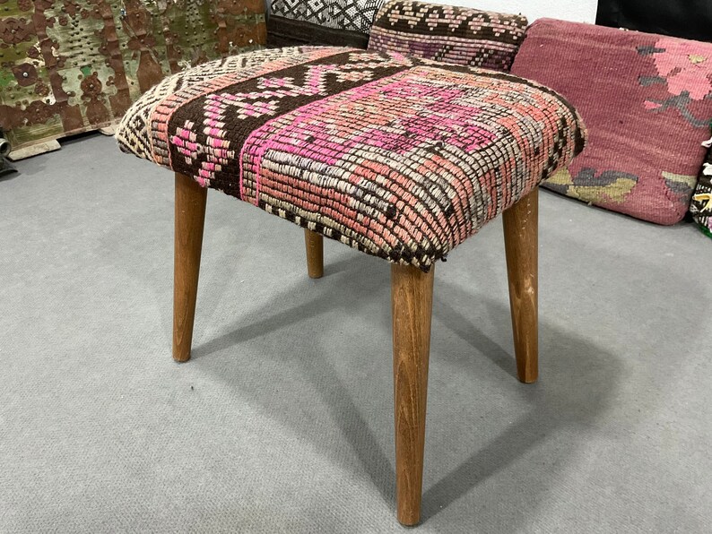 May include: A small, square stool with a patterned top in shades of pink, brown, and cream. The seat is supported by four light-colored wooden legs. The stool has a vintage or handmade aesthetic.